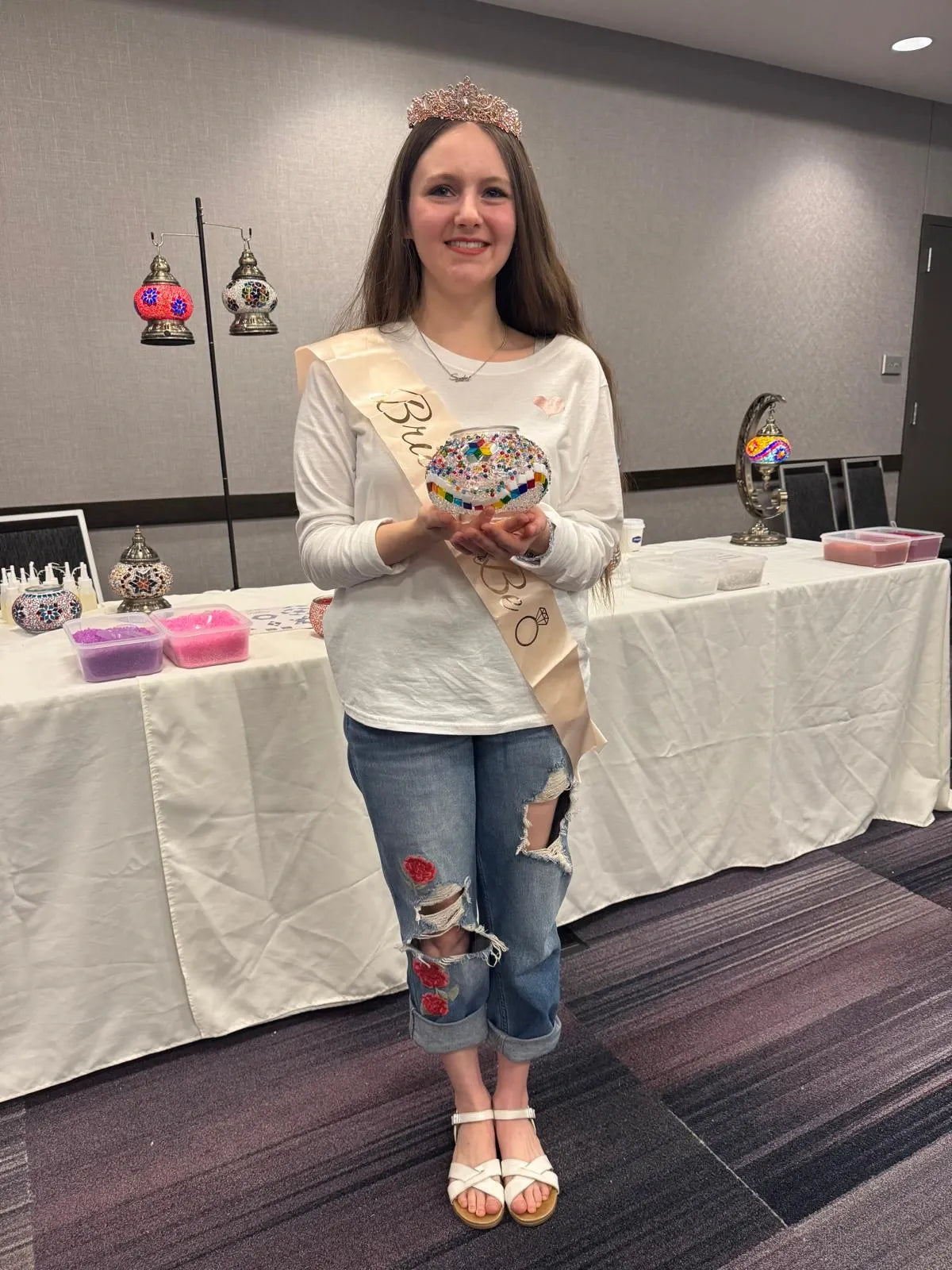 Woman wearing a sash and crown in an indoor setting with tables and decorative items.