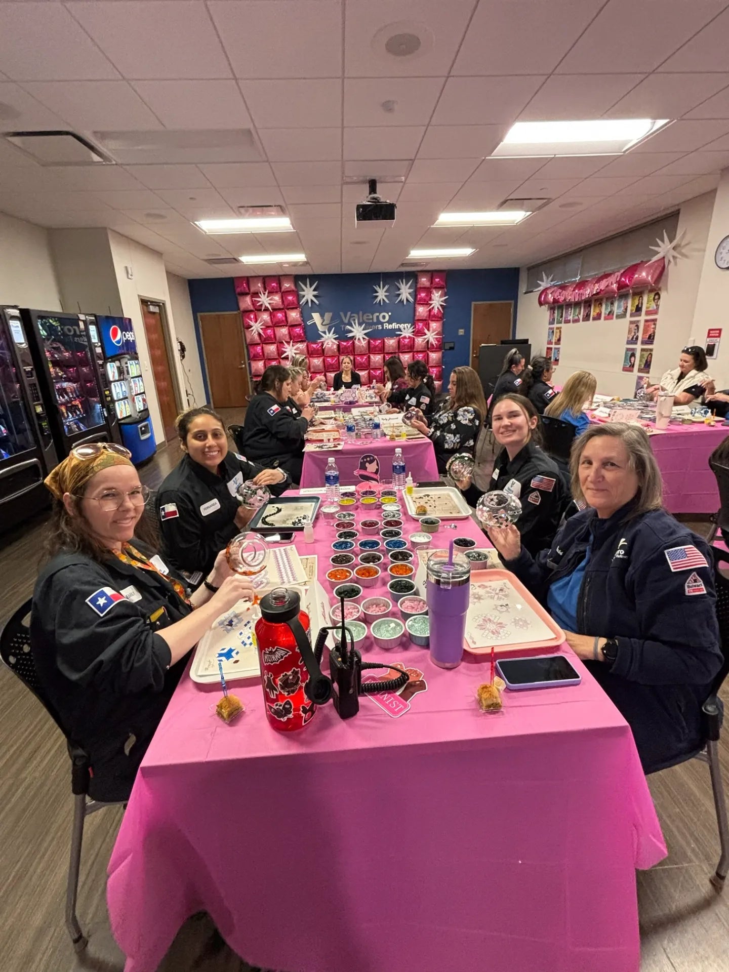 Group of people sitting at a table with pink tablecloth in a room with balloons and decorations.