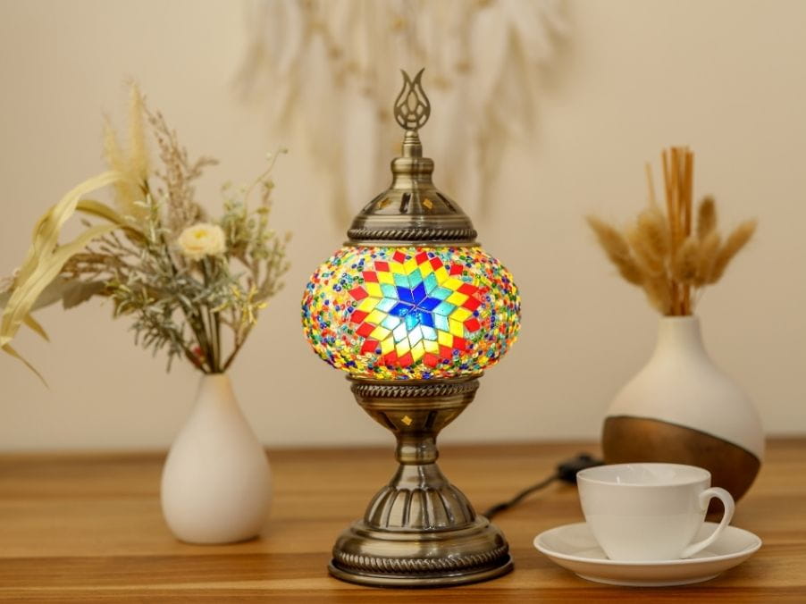 Decorative mosaic table lamp on a wooden surface with a cup and saucer, and vases in the background.