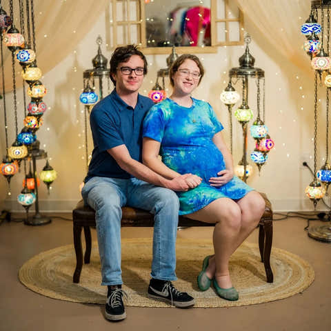 Two people sitting on a chair in a room with colorful hanging lights and a mirror.