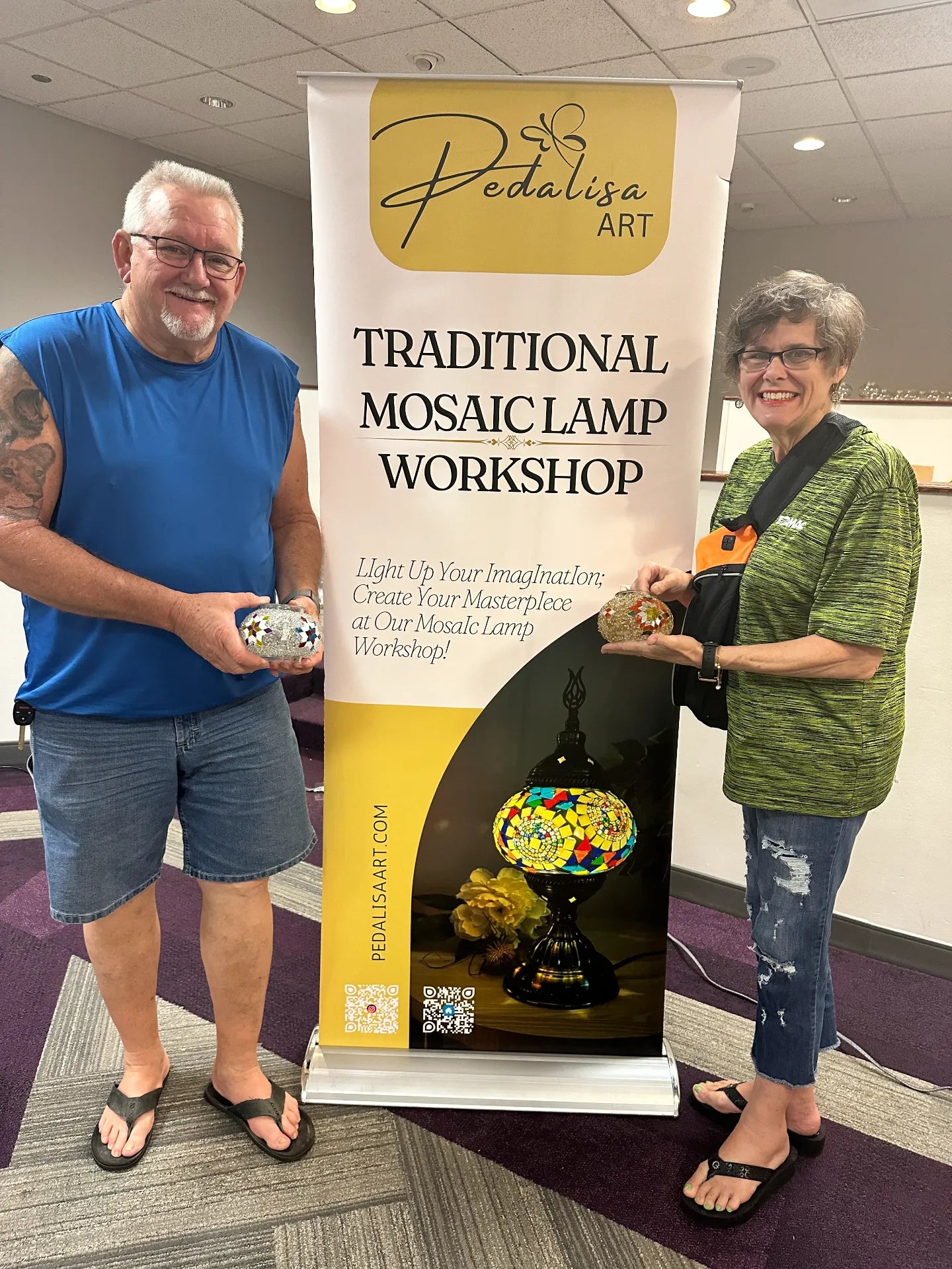Two people holding mosaic artworks in front of a Pedalisa Art workshop banner.
