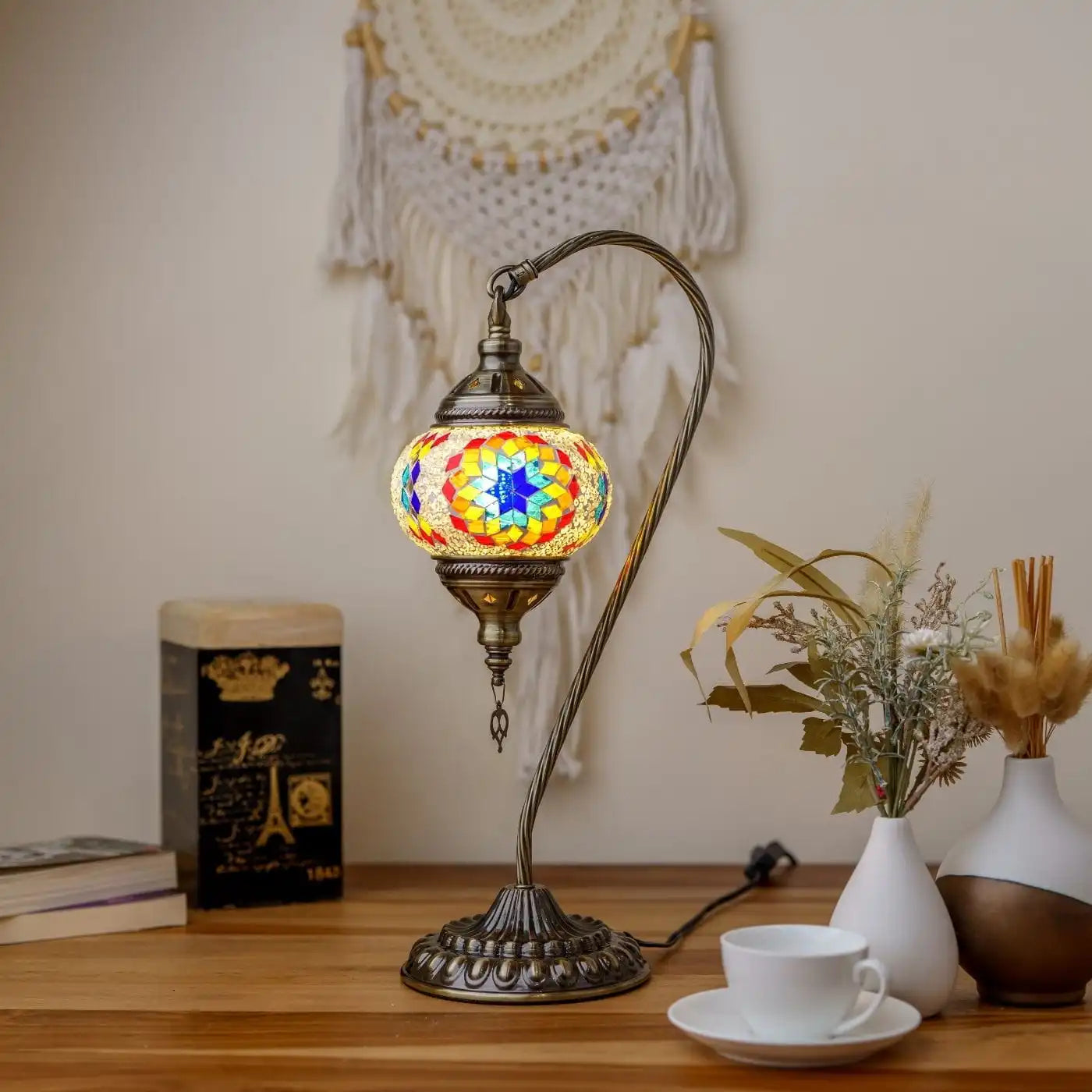 Decorative table lamp with a colorful glass shade on a wooden surface, surrounded by books and a plant.
