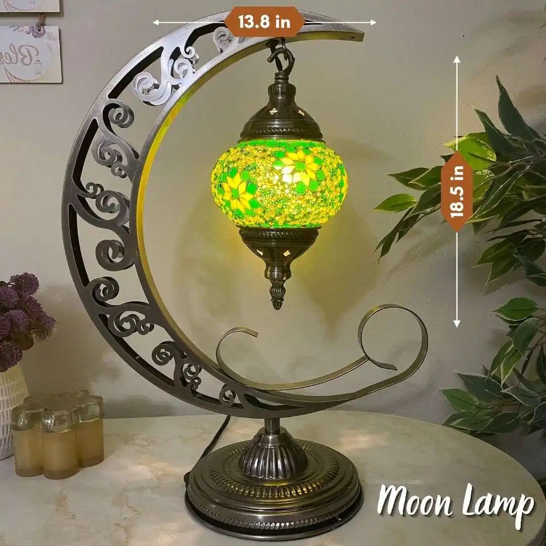 Decorative Turkish Moroccan moon-shaped lamp with a mosaic green and yellow shade, placed on a surface with plants and candles in the background.