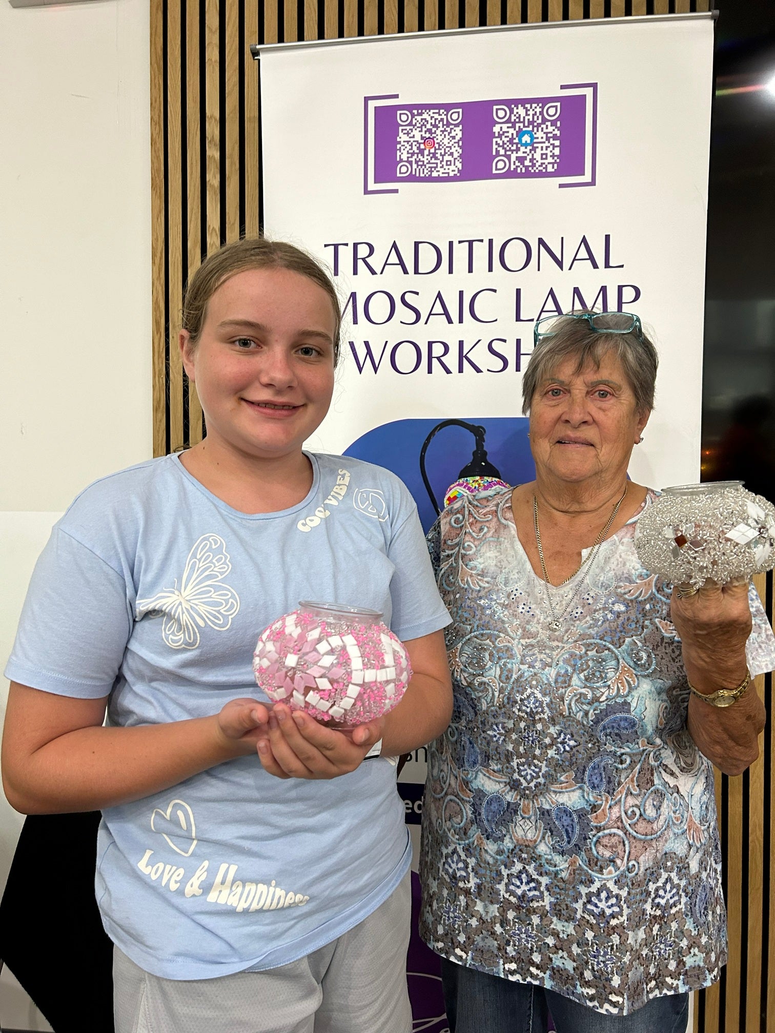 Two women holding mosaic artworks in front of a 'Traditional Mosaic Lamp Workshop' banner.