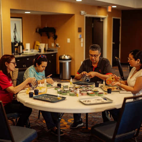 Four people seated around a round table in a hotel-style breakroom or lounge, engaged in conversation and handling food items. The room has warm tan walls, a kitchenette in the background, and blue chairs around the table.