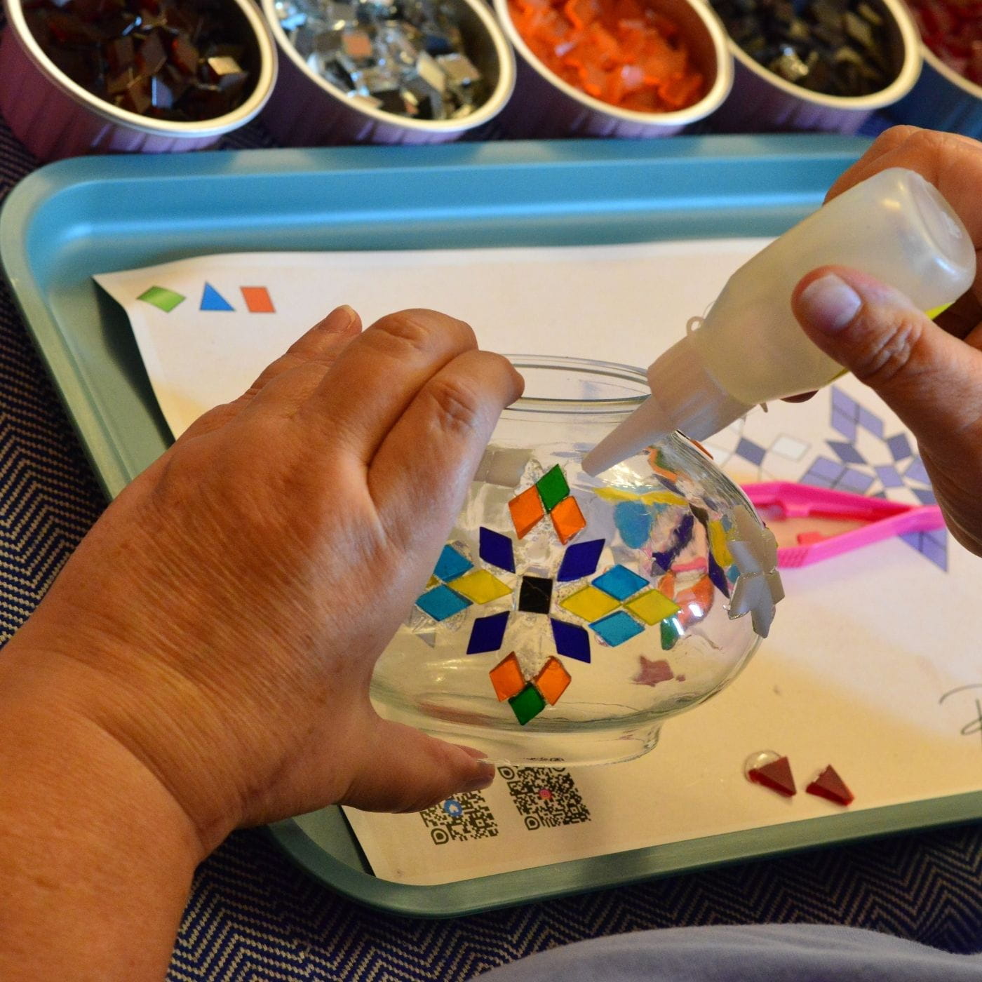 Person adding colorful mosaic pieces to a glass bowl with a tray of materials in the background.