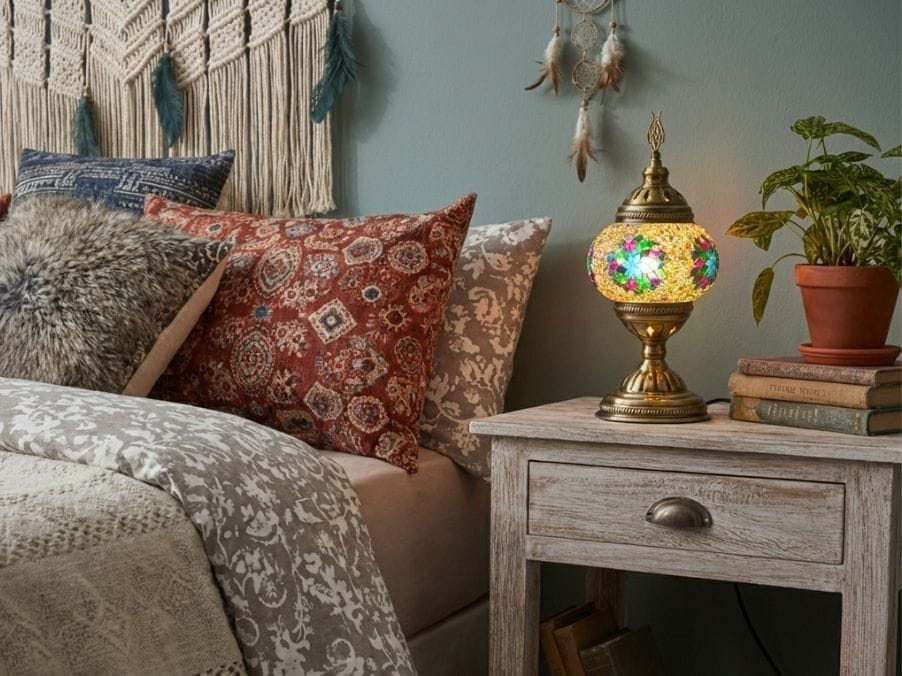 Cozy bedroom with patterned pillows, a decorative mosaic table lamp, and a plant on a wooden nightstand.