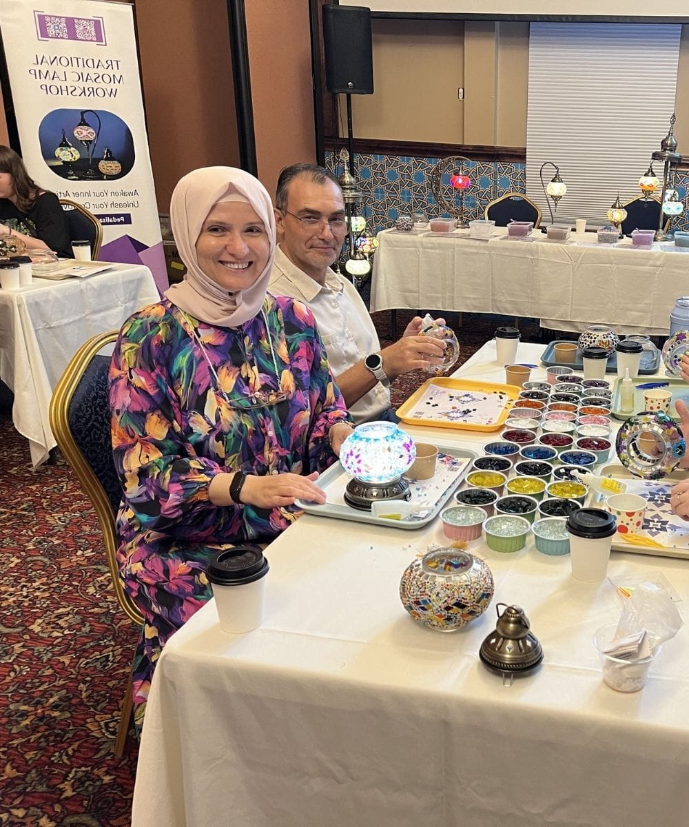 Woman in colorful dress and headscarf sitting at a table with decorative items, smiling.