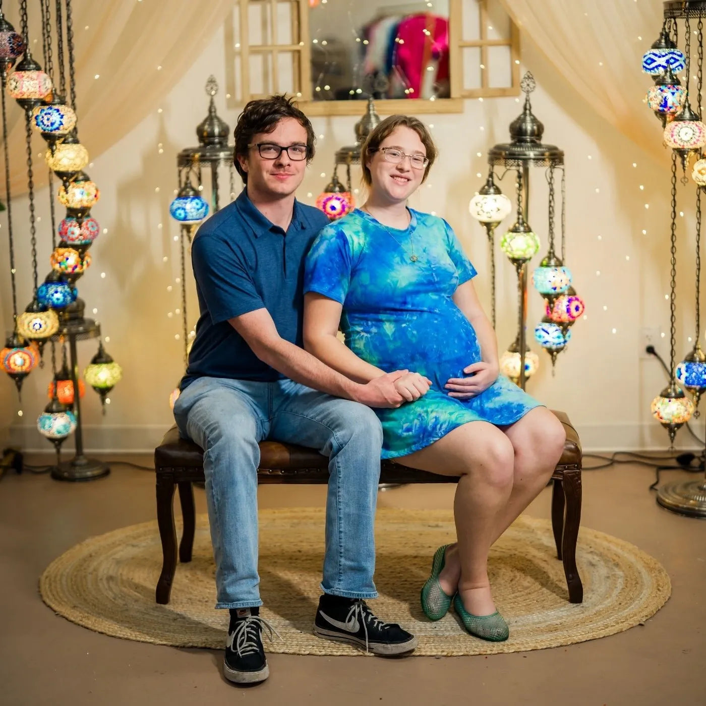 Two people sitting on a chair in a room with colorful hanging lights and a mirror.
