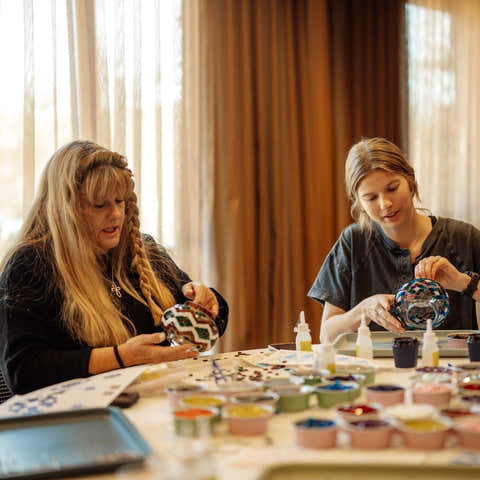 Two women sitting at a table with art supplies, painting ceramic items.
