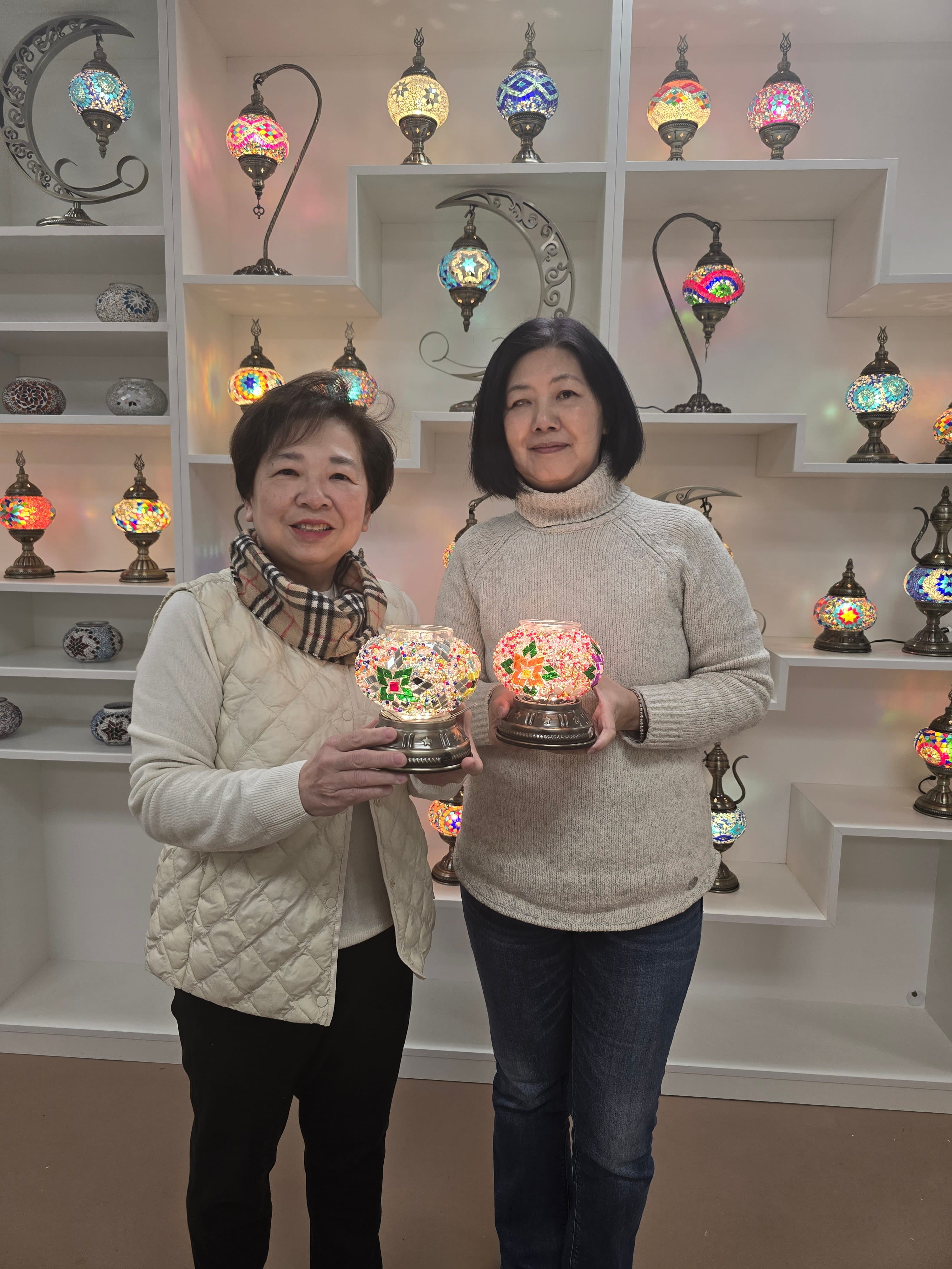 Two women holding decorative lamps in front of a display of similar lamps.