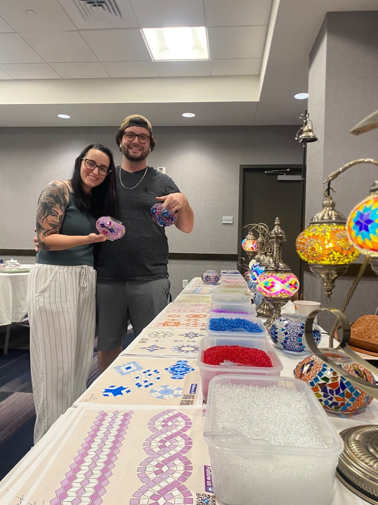 Two people standing behind a table with mosaic art supplies and finished mosaics.