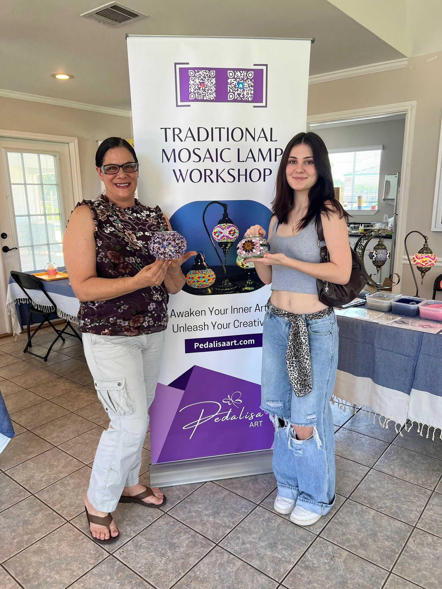 Two women holding mosaic lamps in front of a 'Traditional Mosaic Lamp Workshop' banner.