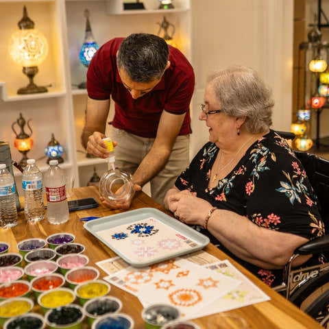 Man assisting an elderly woman with a craft activity at a table.