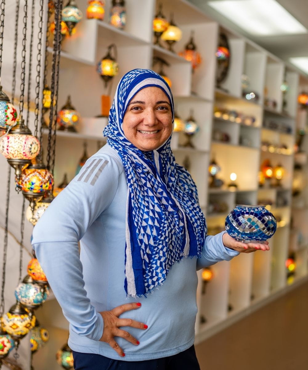 Woman wearing a blue and white headscarf holding a decorative item in a store with hanging lights.