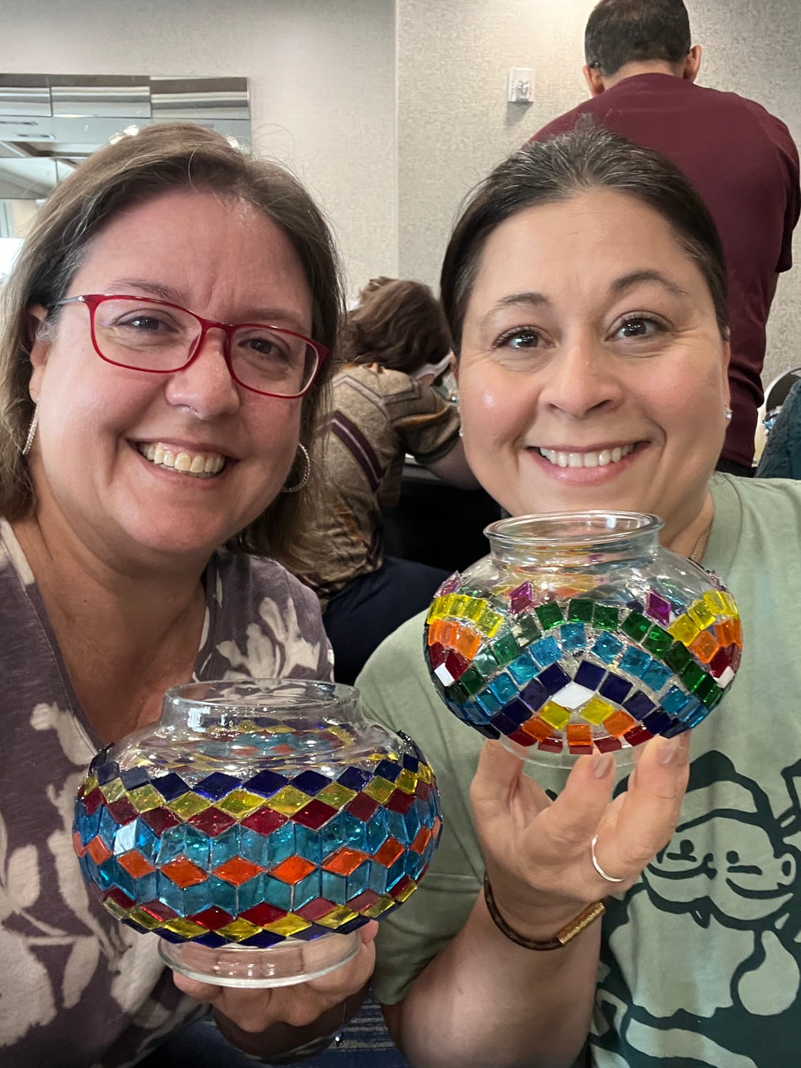 Two women holding colorful mosaic glass bowls in a casual indoor setting.