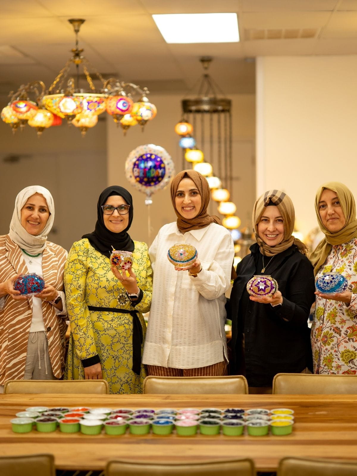 Five women in hijabs standing behind a table with small containers, smiling at the camera.